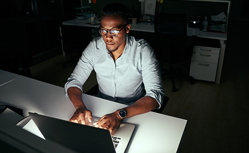 A person wearing glasses works late at night in a dimly lit office, focused on a laptop. The glow from the screen illuminates their face, highlighting a thoughtful expression. The background is dark, with a few faintly visible office furnishings.