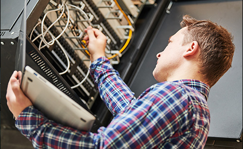 A man in a plaid shirt works on a government network server, holding a laptop in one hand and adjusting cables with the other.