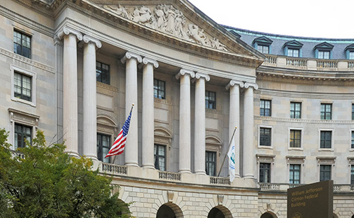 A large, historic building with tall columns and a detailed frieze stands prominently. Two flags, including the U.S. flag, signal its significance in government telecommunications. The curved structure features numerous windows and proudly bears the "William Jefferson Clinton Federal Building" sign.
