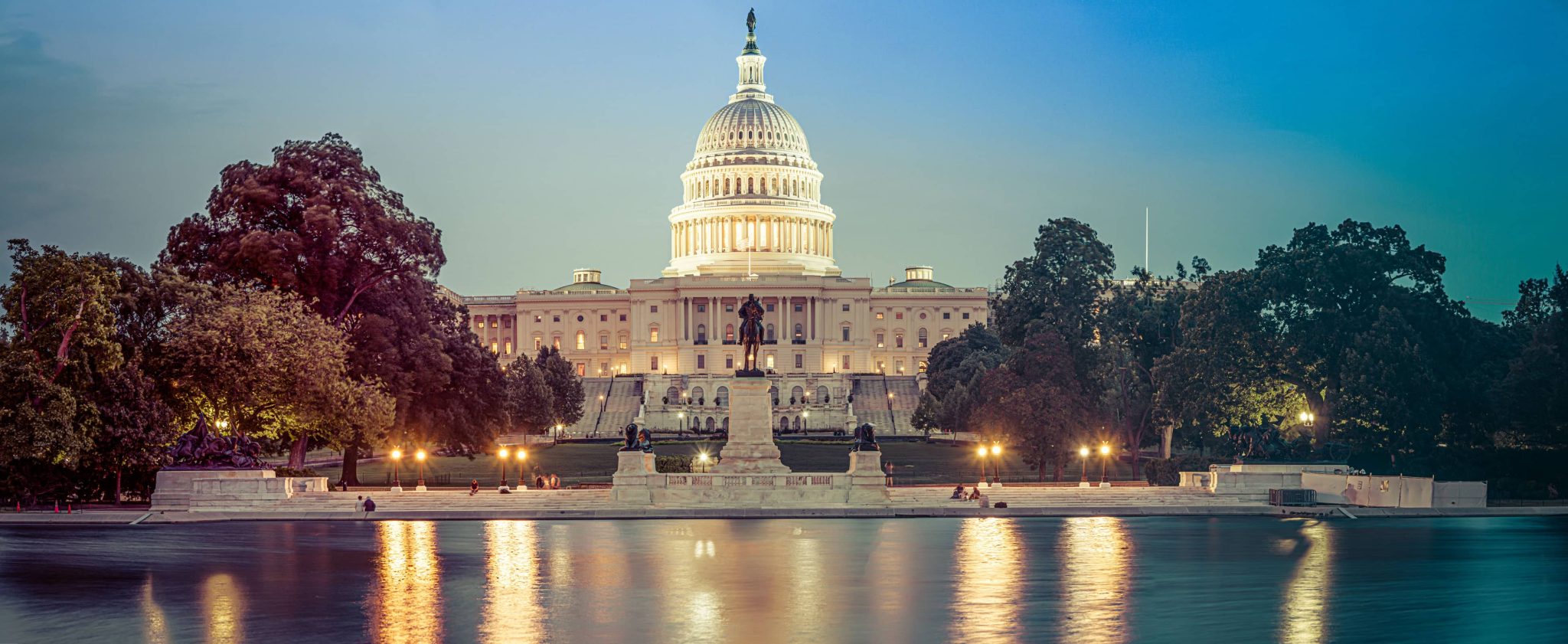 A panoramic view of the U.S. Capitol building at dusk, with its illuminated dome reflecting in a still body of water in the foreground. Framed by trees, this iconic hub of government commands the scene as the sky transitions from blue to pink.
