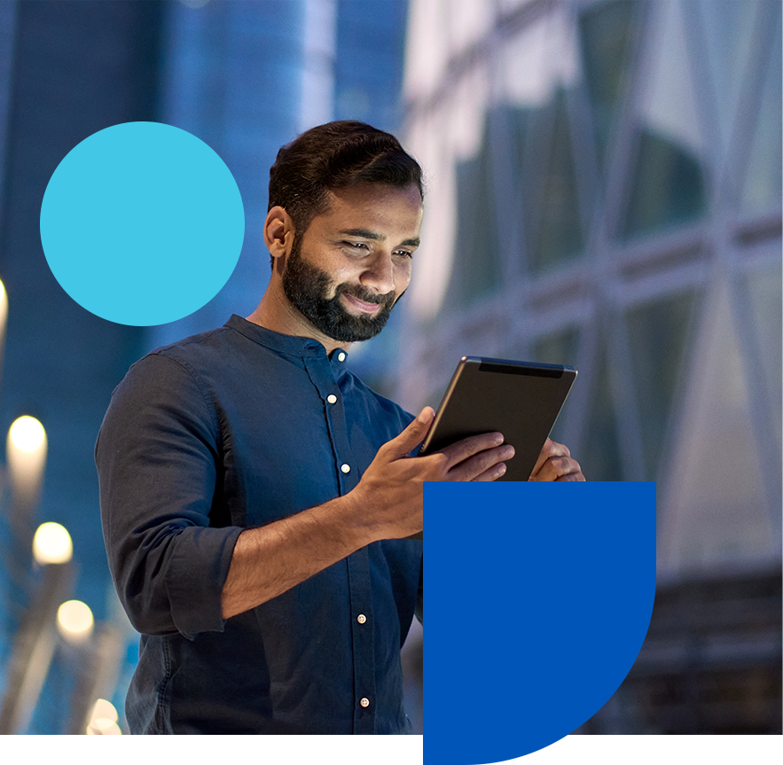 A man in a dark shirt is smiling while using a tablet. He stands in a modern urban setting with blurred lights in the background. Geometric shapes in blue are overlaid on the image.