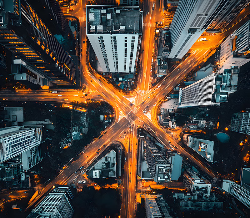 Aerial view of a city at night showing illuminated roads forming an X-shaped intersection, surrounded by tall buildings with glowing windows and vibrant streetlights.