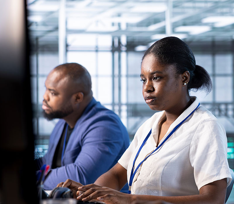 Two professionals sit side by side at desks in an office, focused on their computer screens. The woman in the foreground wears a white shirt and lanyard; the man in the background wears a blue blazer.