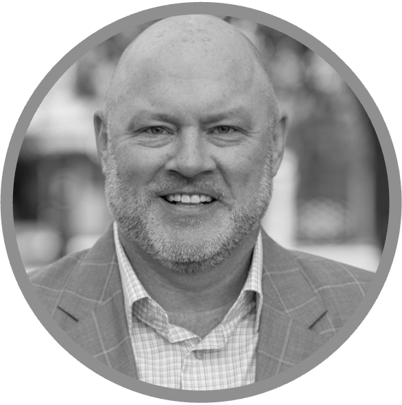 Black and white portrait of a smiling middle-aged man with a bald head and beard, wearing a checked shirt and a plaid suit jacket, inside a circular frame.