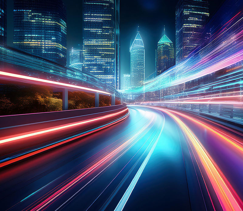 A futuristic cityscape at night with tall illuminated skyscrapers and blurred, colorful light trails from fast-moving vehicles on a curved road, evoking speed, motion, and the pulse of forward thinking telecommunications.