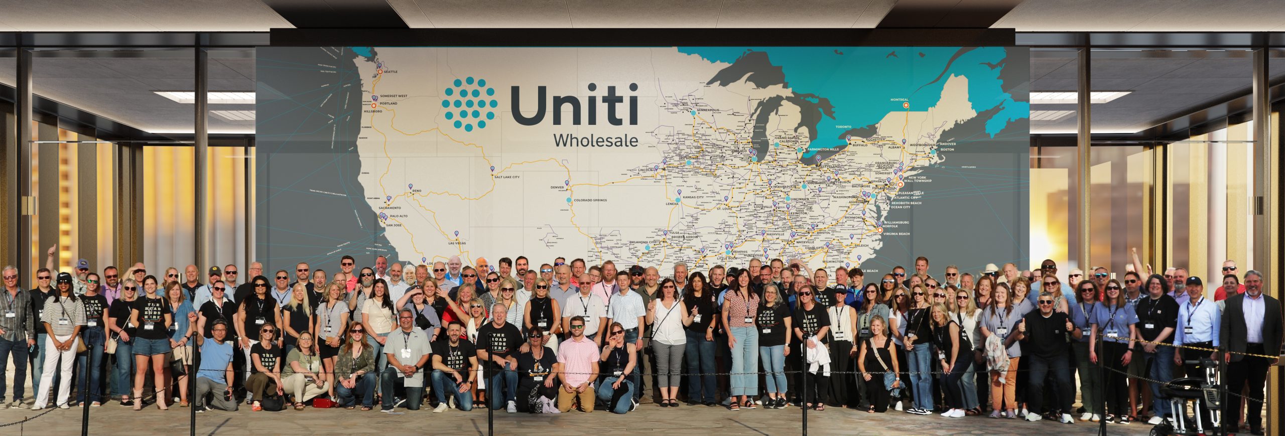 A large group of people pose for a photo in front of a wall-sized United States map labeled “Uniti Wholesale,” showing network routes across the country. The group stands indoors, spanning the width of the map.