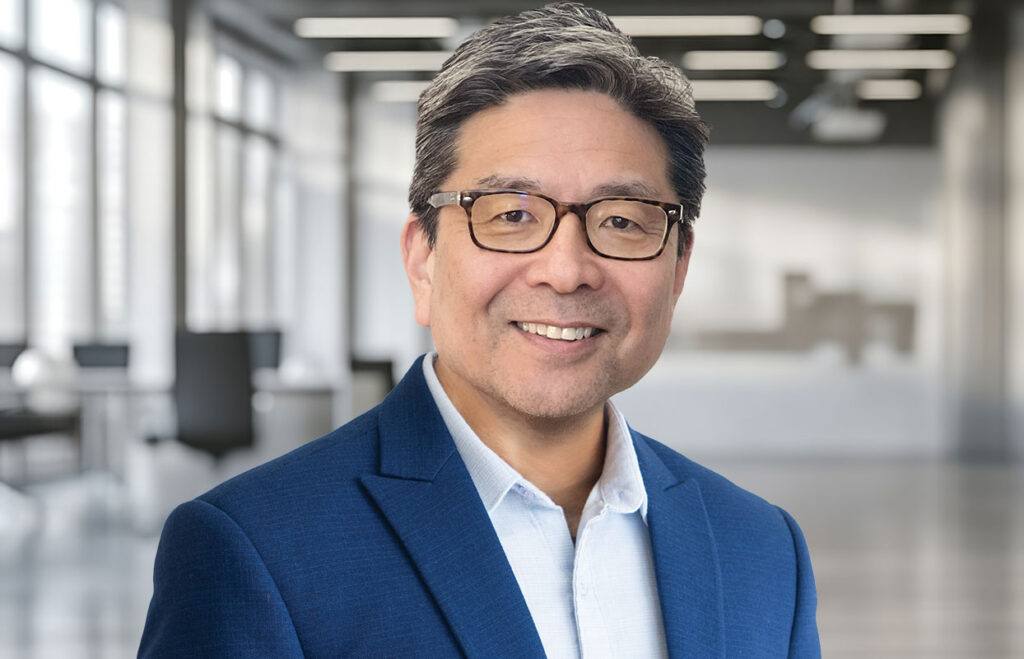 A smiling man wearing glasses, a blue blazer, and a white shirt stands in a modern office with large windows and blurred desks in the background.