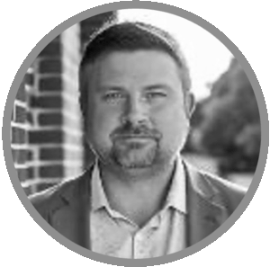 Meet Uniti Wholesale: Black and white portrait of a man with short hair and a trimmed beard, wearing a collared shirt and blazer, standing in front of a brick wall with blurred trees in the background.