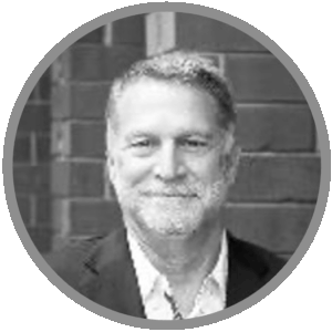 Meet Uniti Wholesale: Black and white portrait of a middle-aged man with short hair and a beard, wearing a suit jacket and open-collared shirt, smiling in front of a brick wall background.