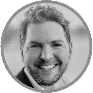 Black and white headshot of a man smiling, with short hair and a beard, wearing a jacket and shirt. The circular image features a gray border and blurred background—Meet Uniti Wholesale’s team in focus.